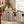 Striped aprons hang on a wooden table with bread, herbs, apples, and copper pots in a sunlit kitchen.