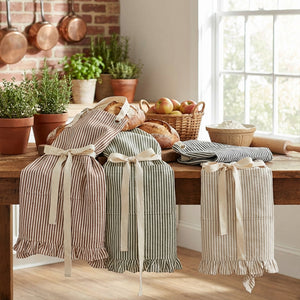 Striped aprons hang on a wooden table with bread, herbs, apples, and copper pots in a sunlit kitchen.