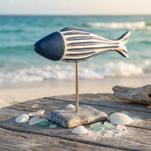 Blue and white fish sculpture on a wooden table, surrounded by seashells, near a sandy beach.