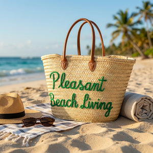 Straw beach bag with "Pleasant Beach Living" text, hat, and sunglasses on sand near ocean.