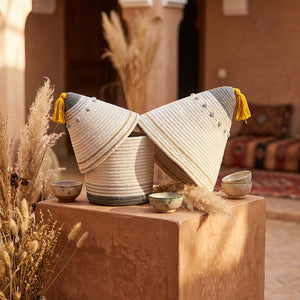 Two woven baskets with yellow tassels, atop a clay pedestal, surrounded by pampas grass.