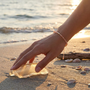 Hand with pink bracelet touching clear glass on sandy beach with seashells, sunset in background.
