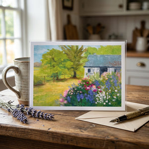 A colorful cottage painting with trees and flowers, displayed on a wooden table with a pen and envelope.