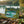 Painting of boats on a turquoise lake, surrounded by lush green trees, placed on a mossy log.