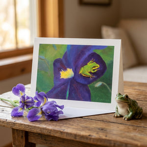A card with a painting of a green frog on a purple flower sits on a table with iris flowers.