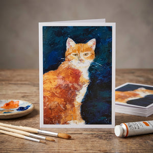 Painting of an orange and white cat on a card, with brushes and paint tube on a wooden table.