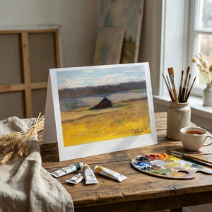 Painting of a barn on a field, surrounded by art supplies on a wooden table near a window.