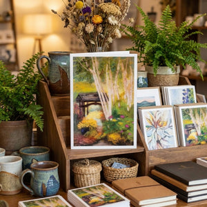 Art display with vibrant landscape painting, surrounded by plants, mugs, baskets, and notebooks.