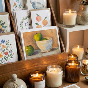 Greeting cards in a wooden display with lit candles nearby; art features flowers and fruit.
