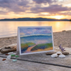 Greeting card with landscape painting on wooden table by lake at sunset, shells and driftwood nearby.
