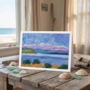 A colorful landscape card on a wooden table with seashells, set in a room with a seaside view.