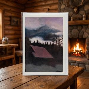 A painting of a cabin in a forest under a moody sky, set on a wooden table by a glowing fireplace.