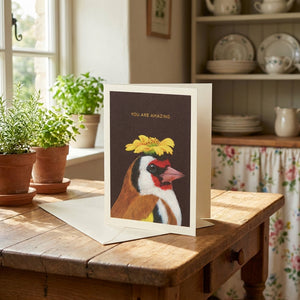Greeting card with a bird and yellow flower, text "You Are Amazing," on a wooden table near herbs.