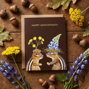 Chipmunks with flowers on a brown anniversary card, surrounded by acorns and leaves on wood.