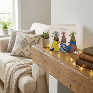 Party card with birds in hats on a rustic mantel, surrounded by string lights and houseplants.