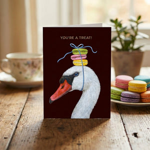Card with a swan in a bow balancing macarons, text "You're a treat!". Macarons and plants nearby.