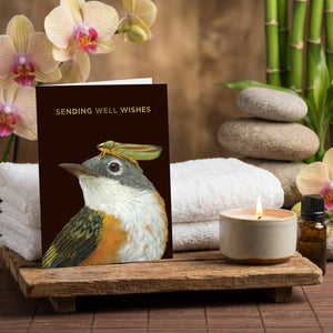 Card with bird and bug illustration, beside orchids, candles, stacked stones, and towels.