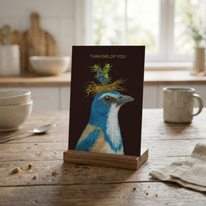 Card with a blue bird and nest on its head, on a wooden table with cups and bowls in the background.