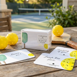 Notes and pickleball items on a wooden table near a court. Bright yellow balls and vibrant cards.