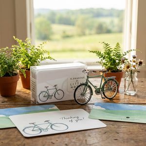 Mini green bicycle, note cards, and potted plants on a wooden table by a window with a countryside view.