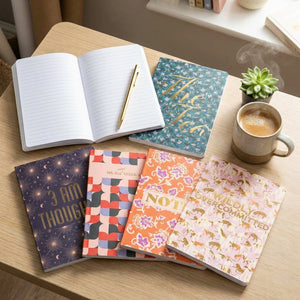Notebooks with colorful covers on a desk, an open lined notebook with a pen, coffee, and a plant.