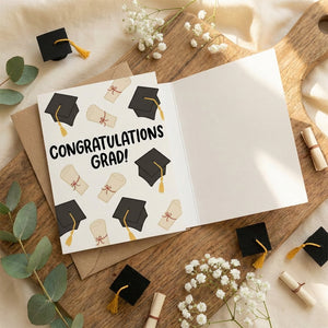 Graduation card with caps and diplomas on wooden board, surrounded by flowers and leaves.