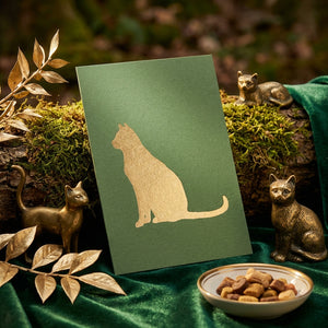 Green card with gold cat silhouette, surrounded by gold cat statues, moss, and a dish of treats.