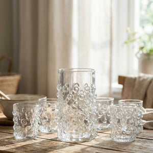 Clear glasses with bubble texture on a wooden table, softly lit by sunlight through a window.