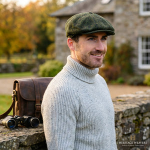 Man in a gray sweater and green cap smiling outdoors, with a leather bag and binoculars nearby.
