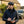Man in a flat cap and quilted vest, writing in a notebook, stands by a rustic house and SUV.