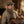 Man in tweed cap and scarf holding leather bag by fireplace; rustic stone wall in background.