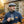 Man in a blue sweater and flat cap holds steaming coffee outdoors near stone building.