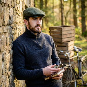 Man in a dark sweater and cap, holding a compass and notebook, leans on stone wall; bicycle nearby.