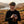 Man in a flat cap holds a vintage camera, standing in a scenic landscape with hills and a lake.