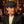 Man in dark blazer and flat cap in a cozy, warmly lit library with leather chairs and bookshelves.