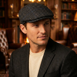 Man in dark blazer and flat cap in a cozy, warmly lit library with leather chairs and bookshelves.