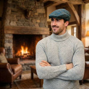 Man in gray sweater and flat cap smiling in rustic room with a stone fireplace and leather chair.