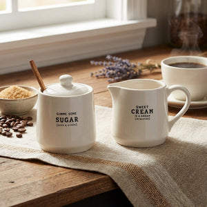 White sugar pot and cream pitcher on a wooden table with coffee beans and a steaming cup.