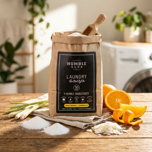 Brown paper bag of laundry soap on wood table with oranges, lemongrass, and laundry in background.