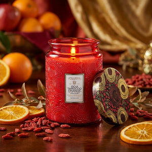 Red jar candle with intricate design, surrounded by dried oranges and goji berries on a wooden table.