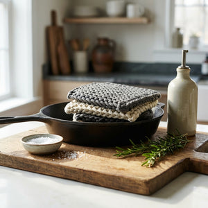 Cast iron skillet with knitted cloths, rosemary, oil bottle, and salt on a wooden board in kitchen.