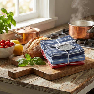 Striped towels, bread, tomatoes, lemons, and herbs on a rustic table near a steaming copper pot.