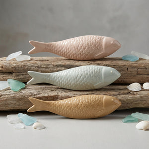 Three ceramic fish in pink, blue, and beige on driftwood with sea glass and shells around.