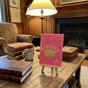 A cozy room with a wooden table, a pink card with a gold crown on an easel, books, and a lamp.