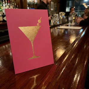 Pink card with a gold martini glass on a wooden bar, with blurred patrons in the background.