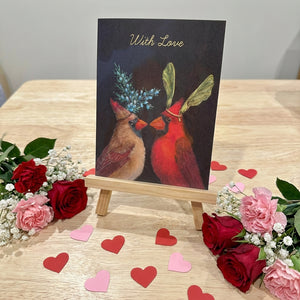 Card with two cardinals on a wooden stand, surrounded by red and pink roses and heart shapes.