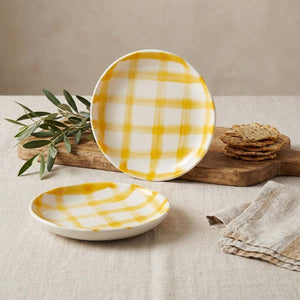 Yellow plaid ceramic plates on a beige tablecloth with crackers and green leaves on a wooden board.
