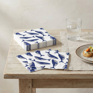 White napkins with blue whale prints on a wooden table, next to a glass of water and a small plate.