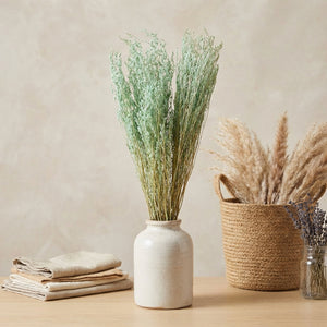 Vase with green foliage, beige basket with pampas grass, and folded linens on a wooden table.