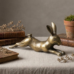 Bronze rabbit sculpture on a linen surface, surrounded by old books and a terracotta plant pot.